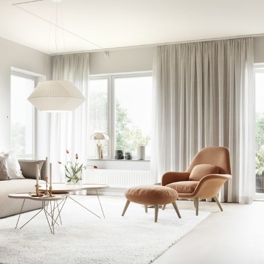 Ljust vardagsrum med stora fönster med designfåtölj med fotpall, soffbord, rislampa, mjuk matta och skira gardiner